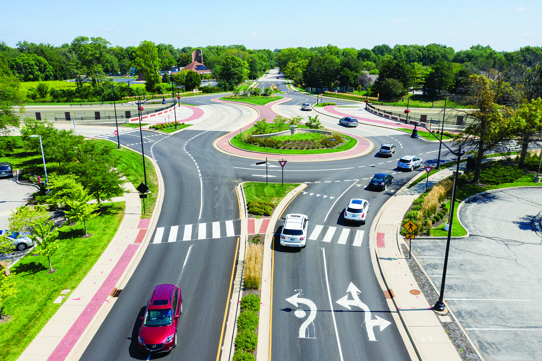 Aerial view of a roundabout with multiple lanes, surrounded by greenery and parked cars. Clear blue sky above.